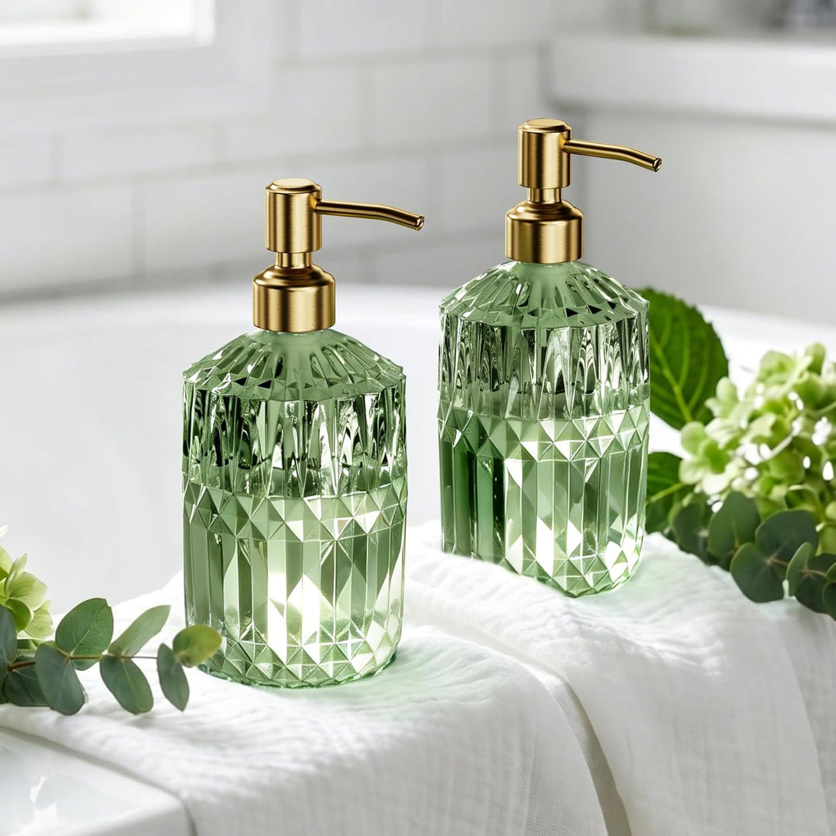 Two green glass soap dispensers with gold pumps on a white surface with greenery.