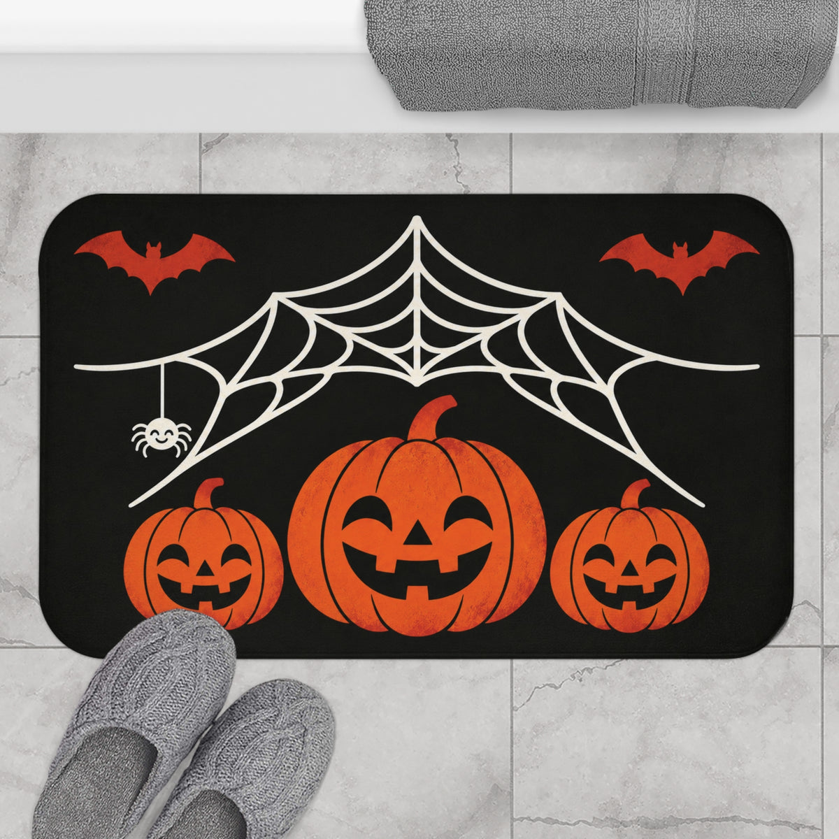 AmazerBath Halloween bath mat featuring spiderweb, bats, and three jack-o'-lanterns placed on light bathroom tile beside a bathtub.