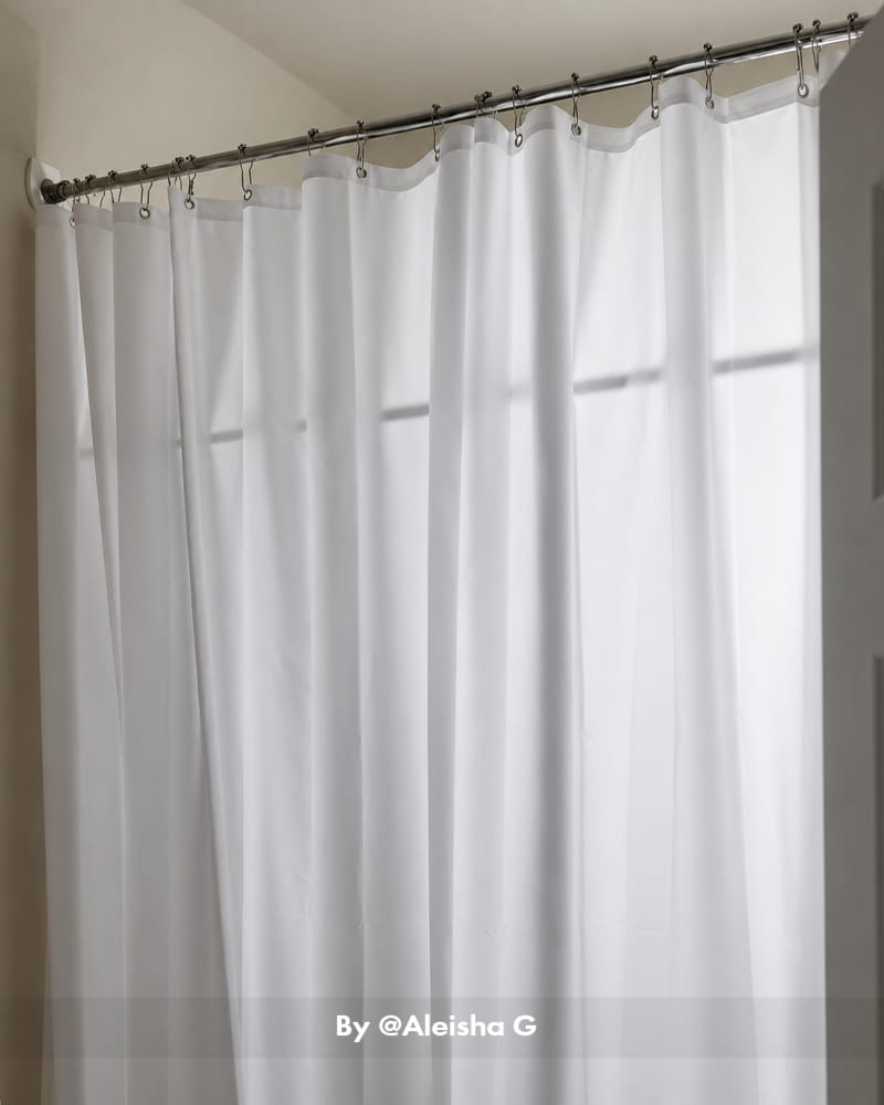 AmazerBath white 120 GSM fabric shower curtain liner softly lit by a window and hanging from chrome shower hooks.