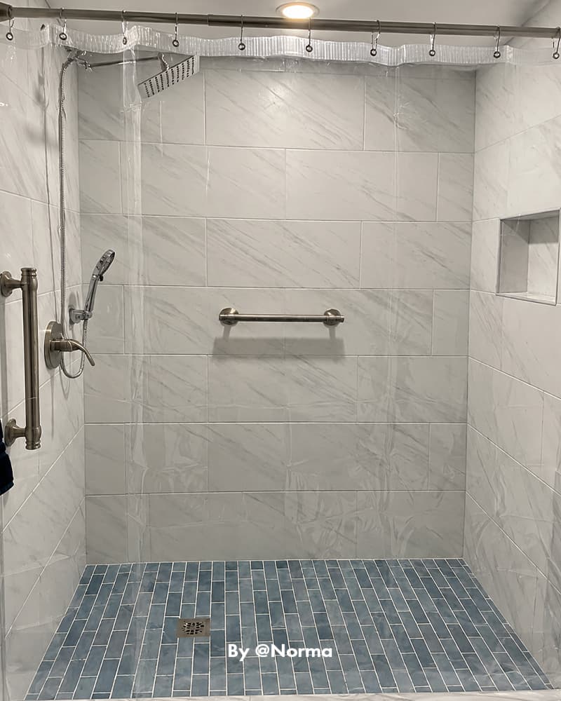 AmazerBath clear 12G PVC shower curtain liner surrounding a marble walk-in shower with grab bars and blue tile flooring.