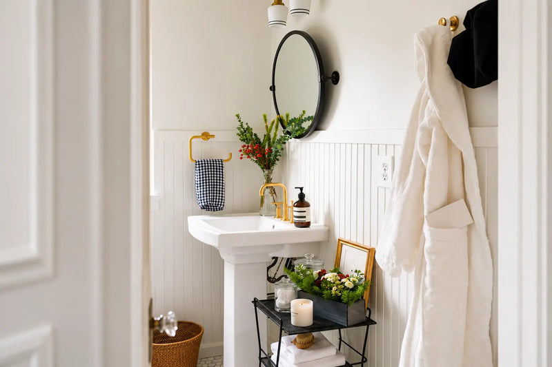 A styled bathroom interior featuring a white pedestal sink with a gold faucet, a round black-framed mirror, a white robe hanging on a gold hook, and decorative plants and accessories.