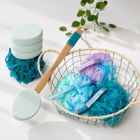 Basket with blue and purple loofahs, a wooden lotion applicator for back, and greenery on a white surface