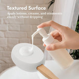 Hand using a white pump bottle to apply cream onto a textured surface.