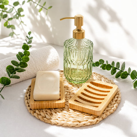 Green glass soap dispenser with gold pump, bar of soap, and wooden soap dish on a woven mat with plants in the background.