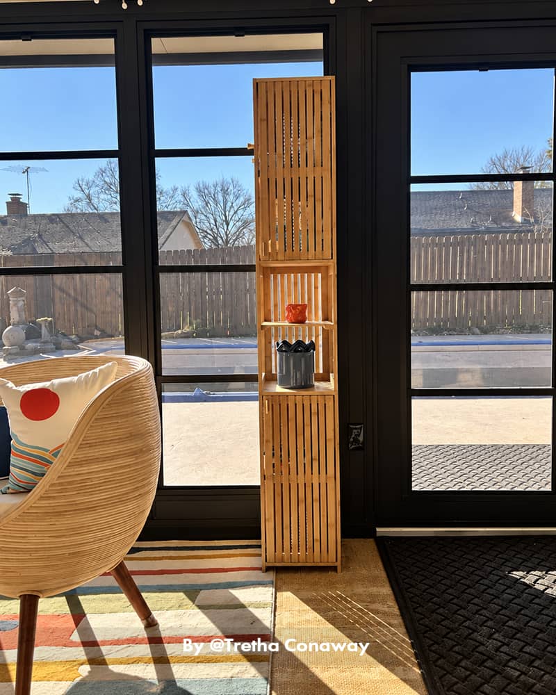 AmazerBath slim bamboo storage cabinet standing by glass patio doors with personal items arranged on its open shelves.