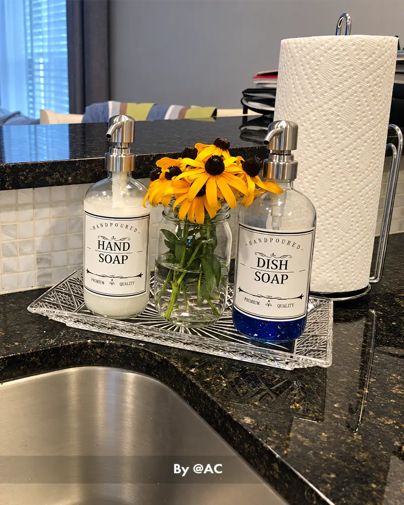 AmazerBath clear glass soap dispensers for hand and dish soap displayed by a kitchen sink with flowers and paper towels.
