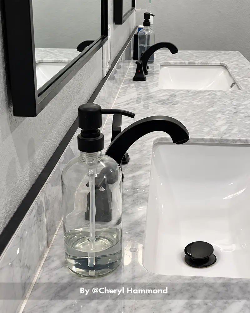 AmazerBath clear glass soap dispenser with a matte black pump placed beside a black faucet on a marble bathroom vanity.