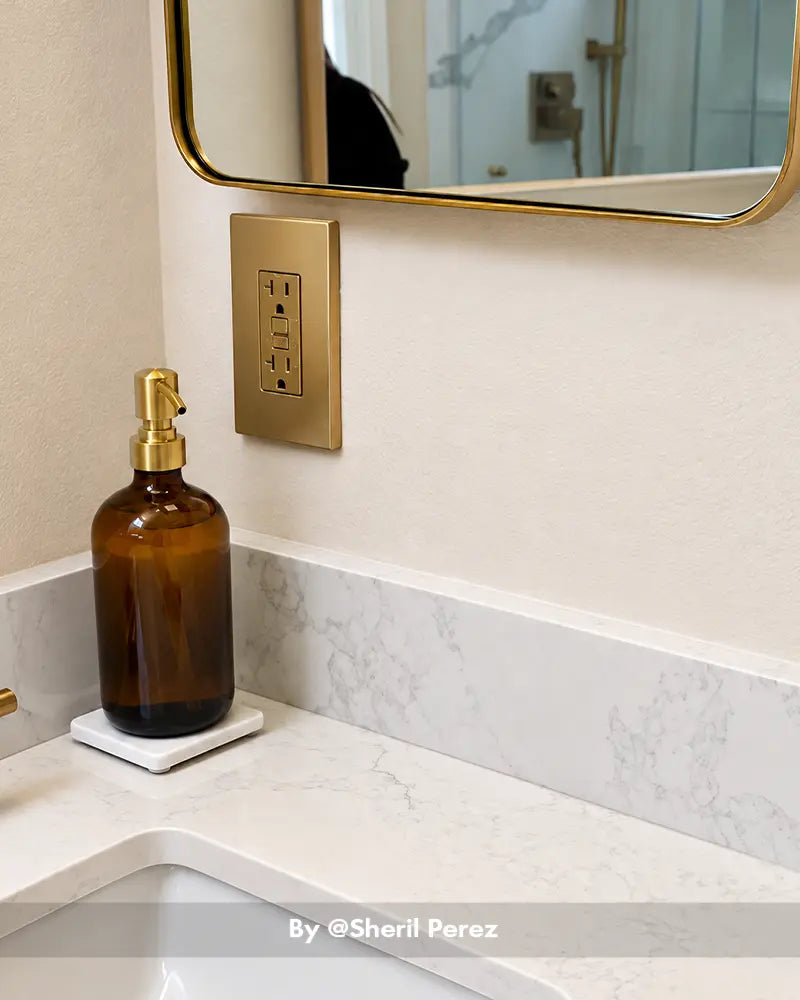 AmazerBath amber glass soap dispenser with a gold pump resting on a white tray beside a marble bathroom sink and gold mirror.