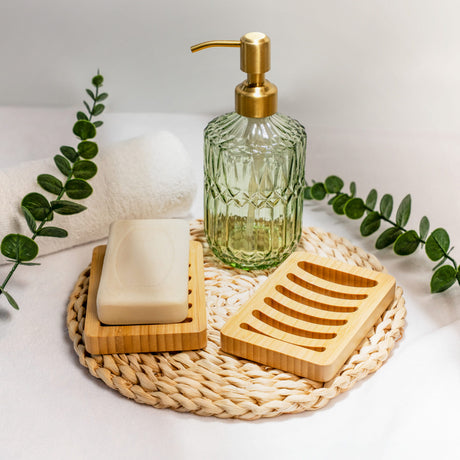 Glass soap dispenser with gold pump, soap bar, and wooden soap dish on a woven mat with green leaves.