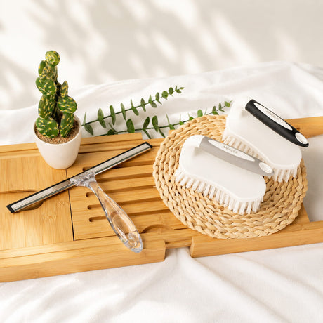 Cleaning tools including a squeegee, scrub brush, and white container on a wooden tray with a plant in the background.