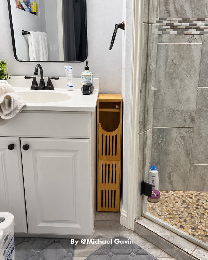 AmazerBath bamboo toilet paper storage cabinet placed between a white vanity and tiled walk-in shower.