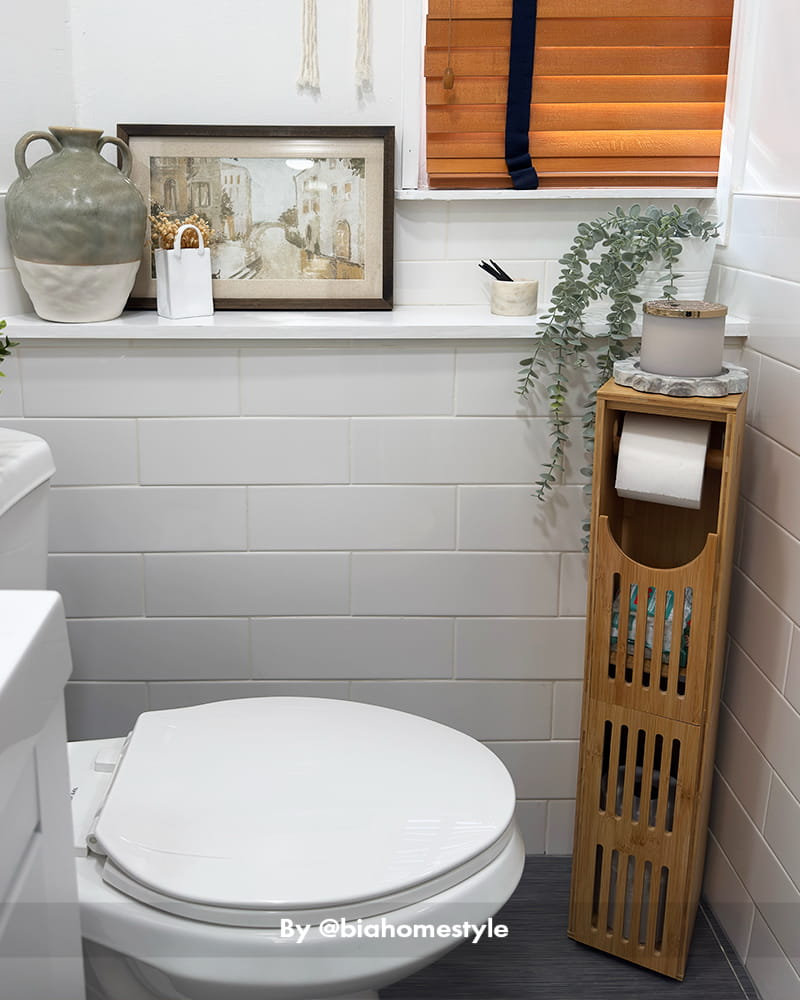 AmazerBath bamboo toilet paper storage cabinet holding rolls beside a white toilet with plants and framed decor.