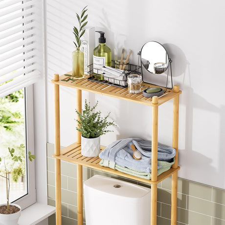 AmazerBath Natural bamboo two-tier over-the-toilet shelf holding toiletries, folded towels, a mirror, and a plant above a toilet.